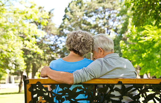 Retired man sitting with arm around woman at nursing home. Rear view of senior couple is relaxing on bench at back yard. They are spending leisure time.