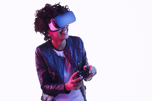 Lovely African American teen girl in VR goggles smiling and using controller while playing game on white background