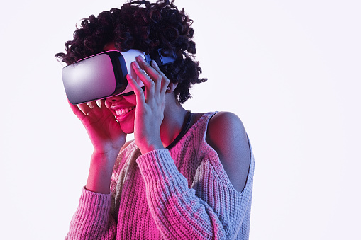 Pretty African American teen girl smiling and looking away while wearing futuristic VR headset and standing on white background