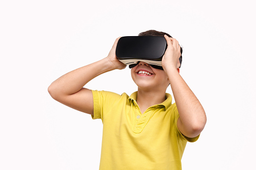 Cute boy in yellow T-shirt smiling and wearing modern VR headset while standing on white background