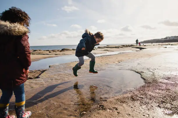 Little boy wearing wellington boots jumping and splashing in a puddle of water on the beach.