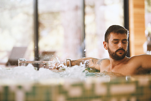 A man is resting in a hot tub in a wellness resort. He closed his eyes and enjoys the sun shining through the windows.