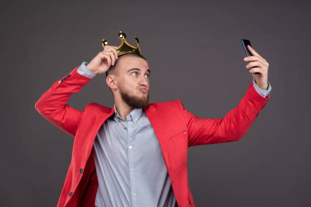 junge bärtige hübsche mann in goldkrone nimmt selfie mit blick auf smartphone - arroganz stock-fotos und bilder