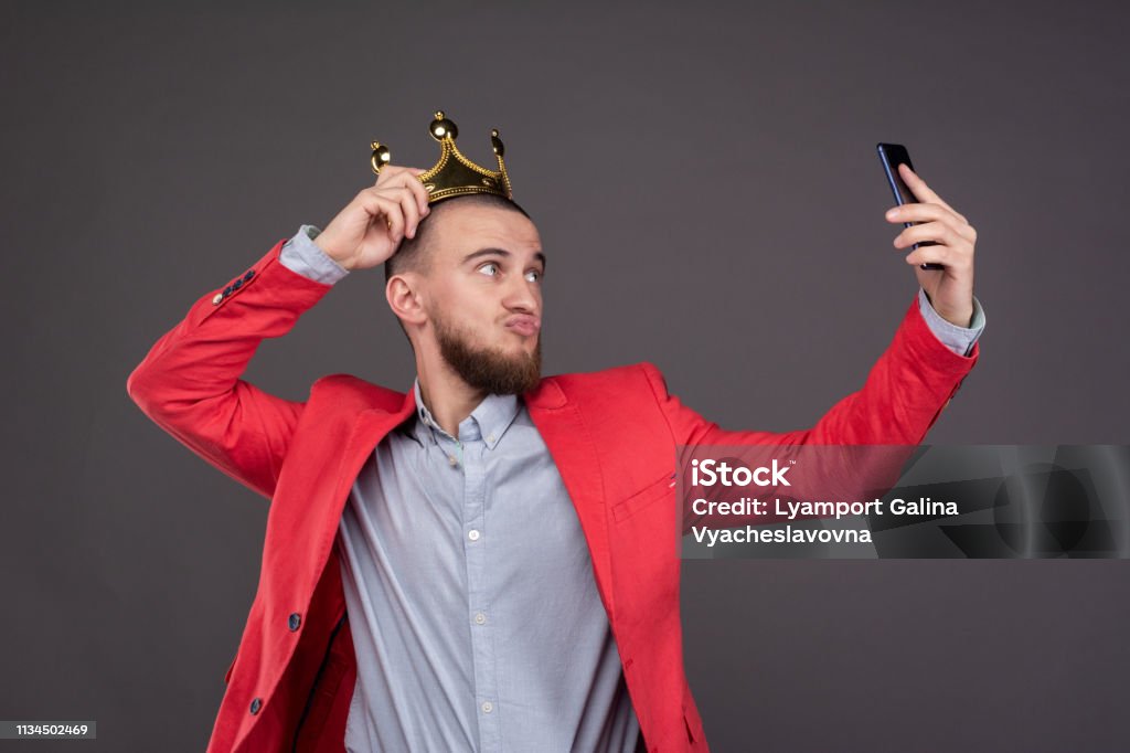 Young bearded handsome man in gold crown taking selfie looking at smartphone Vanity Stock Photo