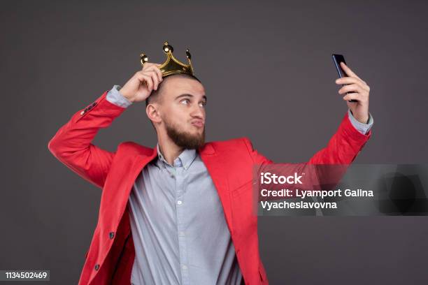 Young Bearded Handsome Man In Gold Crown Taking Selfie Looking At Smartphone Stock Photo - Download Image Now