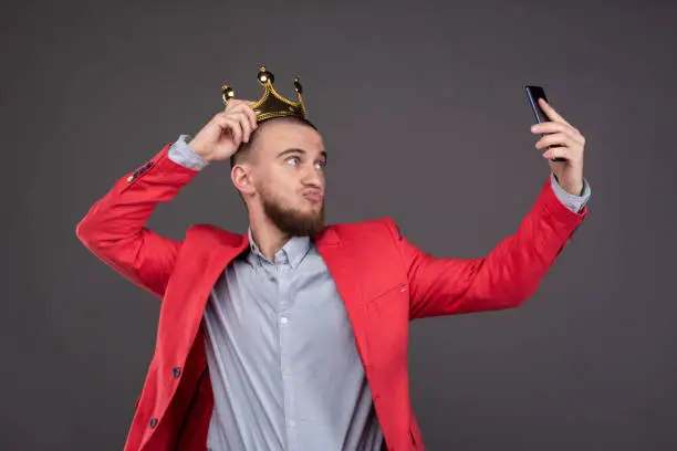 Photo of Young bearded handsome man in gold crown taking selfie looking at smartphone