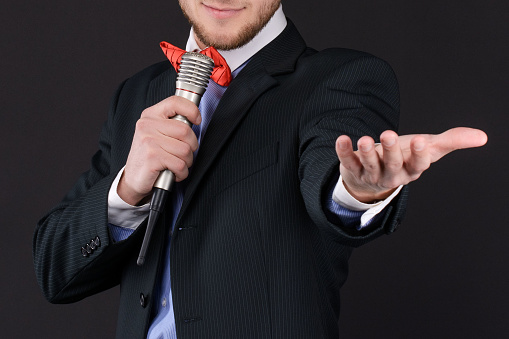 Handsome Master of ceremonies in black suit holding microphone in hand on black background. Showman, tv. closeup