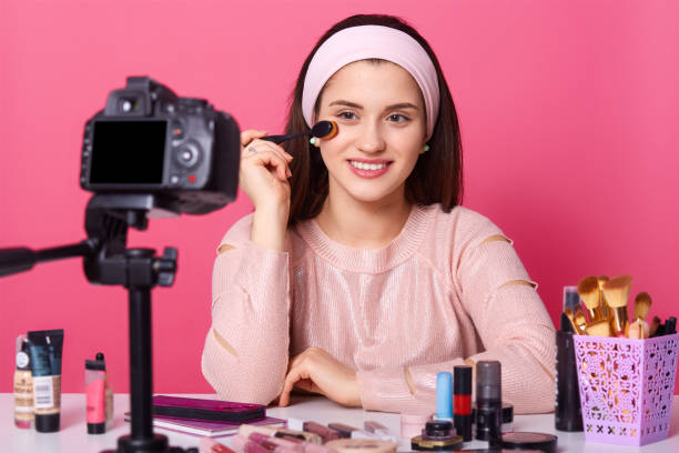 istock Smiling female records make up tutorial video, holds cosmetic brushes. Beautician applies foundation to face in front camera. Vlogger advertises cosmetics isolated over pink wall. Beauty concept. 1134556594
