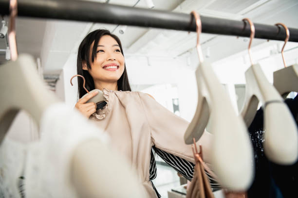 Candid portrait of woman selecting clothing on rail View through coat hangers towards mid adult Chinese woman choosing new clothes in boutique, retail, shopping, choice women satisfaction decisions cheerful stock pictures, royalty-free photos & images