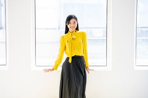 Attractive Chinese woman standing by window, wearing yellow blouse and black skirt, looking at camera, smiling, cheerful, confidence