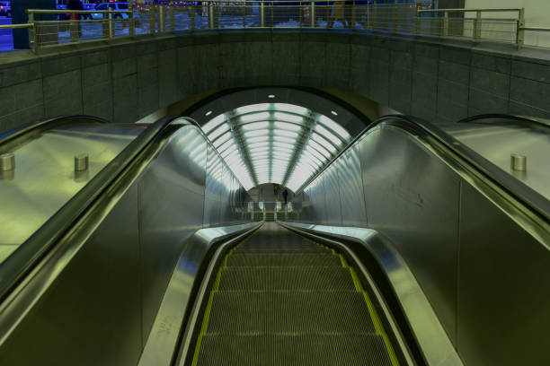 Stock photo showing close-up, rear view of a metal escalator, in an indoor retail area, being ridden by an Indian man wearing a short-sleeved shirt and shorts, and carrying a backpack.\n\n* Not AI modified *