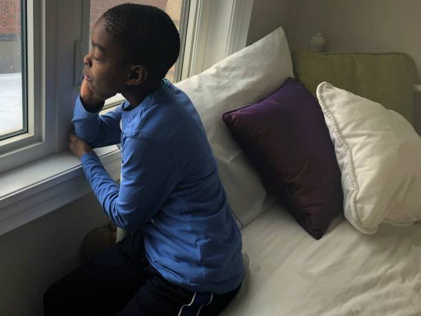 A young boy sitting on a bed looking out the window on a gloomy day