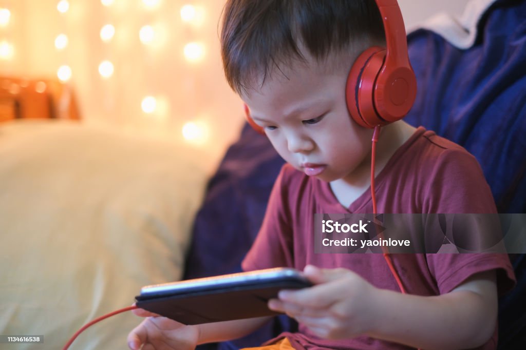 Cute little Asian 2 -3 years old toddler boy child listening to music with headphones from smartphone Cute little Asian 2 -3 years old toddler boy child listening to music with headphones from smartphone, Kids playing with phone in bedroom at home, Gadget-addicted children, internet addiction concept Child Stock Photo