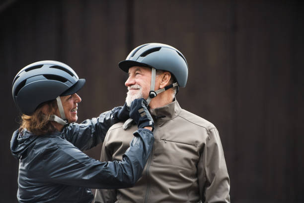 pareja senior activa con cascos de bicicleta de pie al aire libre againts fondo oscuro. - cycling senior adult sports helmet men fotografías e imágenes de stock