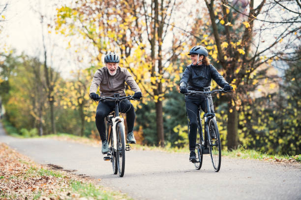 a senior couple with electrobikes cycling outdoors on a road in park in autumn. - senior adult action cycling senior couple imagens e fotografias de stock