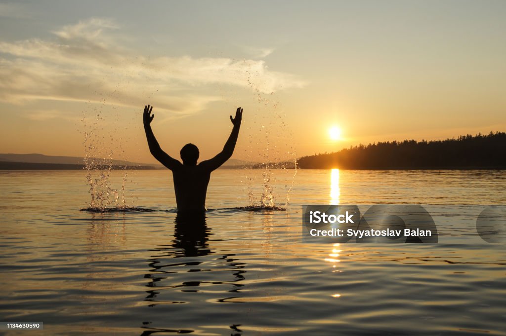 Sagoma di un uomo sullo sfondo del tramonto. Il ragazzo in vita si tuffò nel mare. Spruzza acqua a mano. - Foto stock royalty-free di Acqua