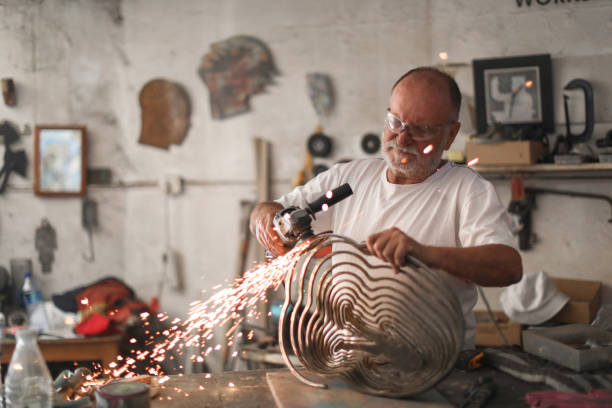 a Senior man creating sculptures in his art studio a Senior man using an angle grinder to create sculptures out of metal in his art studio. sculptor photos stock pictures, royalty-free photos & images