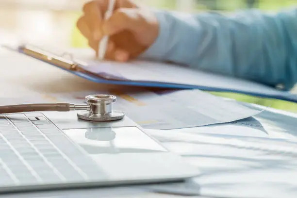 Photo of Doctor working in hospital writing prescription clipboard, working an Laptop on desk in hospital with report analysis, Healthcare and medical concept, selective focus