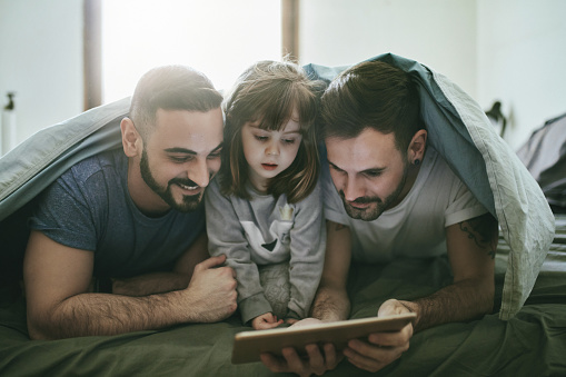 Gay couple looking at digital tablet with daughter in bedroom. Cute girl is watching movie with parents on bed. They are covered with blanket.