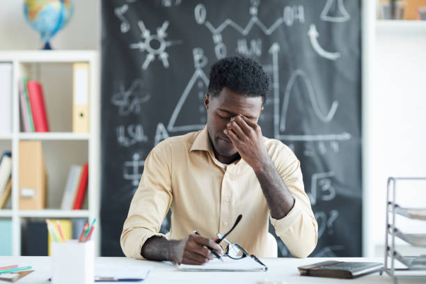 Tired teacher Tired young teacher sitting by desk in classroom and touching nose bridge after several lessons instructor stock pictures, royalty-free photos & images