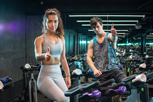 Beautiful sporty young woman and handsome man showing thumbs up while sitting on indoors cycling bikes in the gym