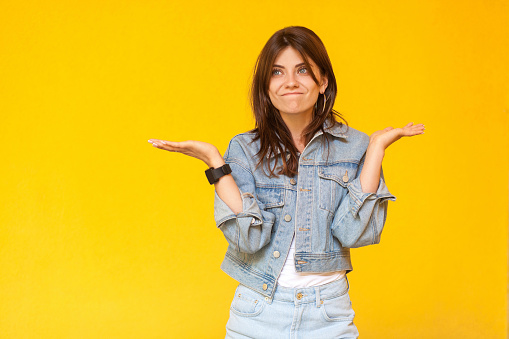 I don't know. Portrait of confused beautiful brunette young woman with makeup in denim casual style standing with raised arms, looking away and doubt. indoor studio shot, isolated on yellow background