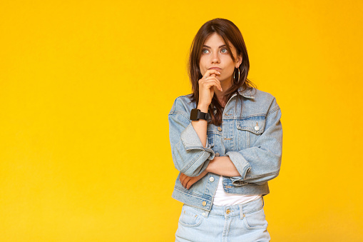 Portrait of thoughtful beautiful brunette young woman with makeup in denim casual style standing, touching her lips, looking away and thinking what to do. studio shot, isolated on yellow background.