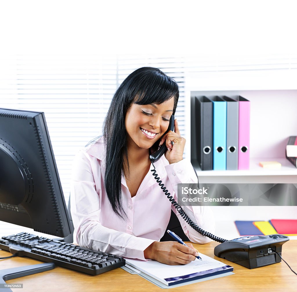 Smiling black businesswoman on phone at desk Smiling young black business woman on phone taking notes in office Telephone Stock Photo