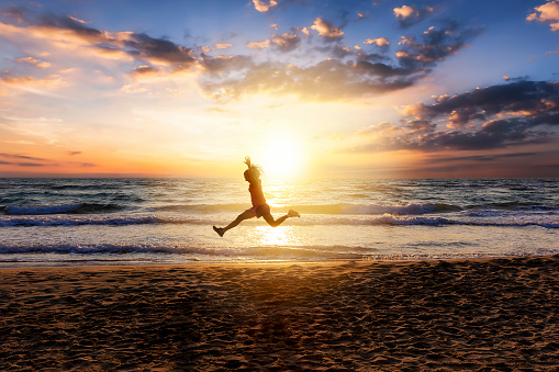 Woman runs and jumps in the air during her outdoor exercise on the beach during sunset time