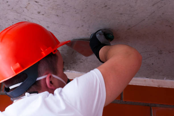 A man in a helmet and respirator performs painting work on a concrete ceiling, against a background of brickwork. The concept DIY Do It Yourself