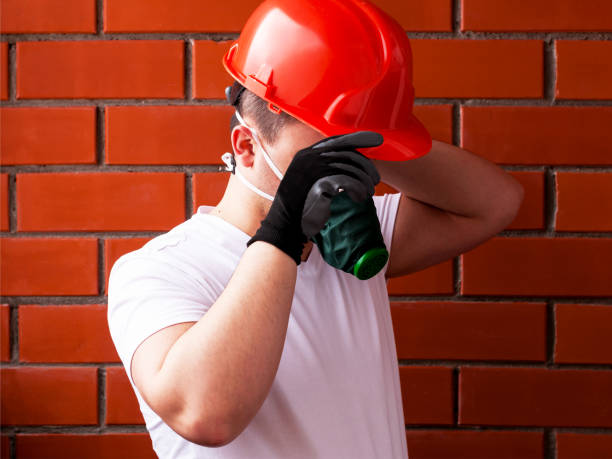 A man in a helmet and respirator performs painting work on a concrete ceiling, against a background of brickwork. The concept is workplace safety. DIY Do It Yourself.