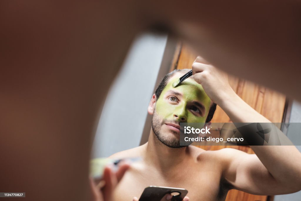 Man Applying Face Mask For Male Beauty In Home Bathroom Latin homosexual person with beard grooming in bathroom at home. Gay metrosexual man applying beauty mask with brush and lotion on face skin Bathroom Stock Photo