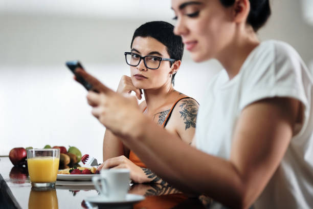 Jealous Lesbian Woman Looking at Partner Messaging On Social Media Domestic lifestyle with two lesbian women at home eating breakfast, partner chatting on mobile telephone. Young woman being ignored by her girlfriend and feeling jealous sad gay stock pictures, royalty-free photos & images