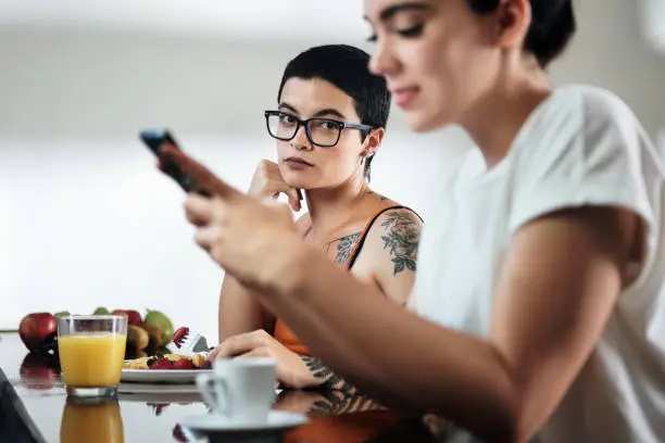 Domestic lifestyle with two lesbian women at home eating breakfast, partner chatting on mobile telephone. Young woman being ignored by her girlfriend and feeling jealous