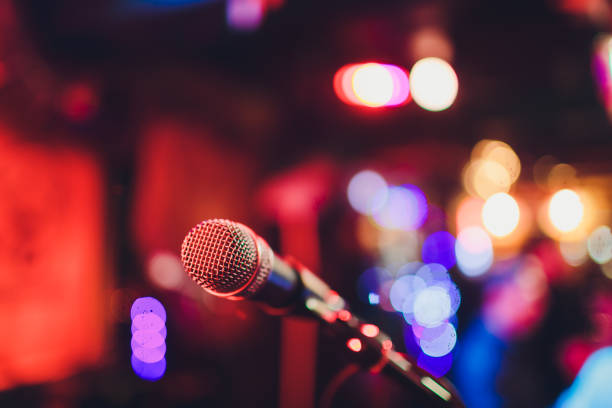 istock Microphone on a stand ready for live music performance or karaoke night with soft bokeh lights and people silhouettes in the background. Concept for musical singing event, having a good time. 1134790092