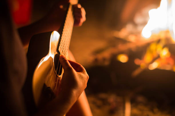 a female musician playing guitar outside, sitting next to a fire. relaxation. - campfire imagens e fotografias de stock