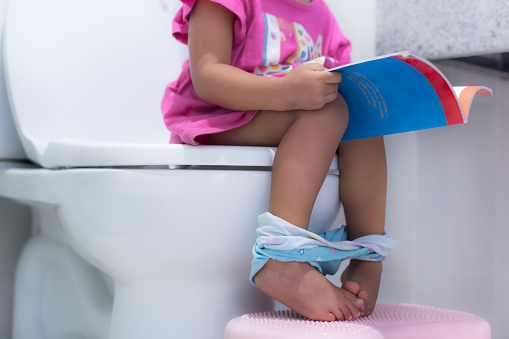 A young female kid sitting on a big toilet in the bathroom reading a book while waiting to go. Close up on legs. Unrecognizable.