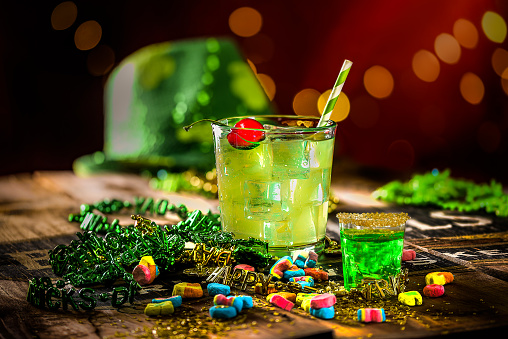 Shamrock Sour cocktail with green gelatin shot and festive St. Patrick's Day party favors and decorations on rustic wood with red background and lights