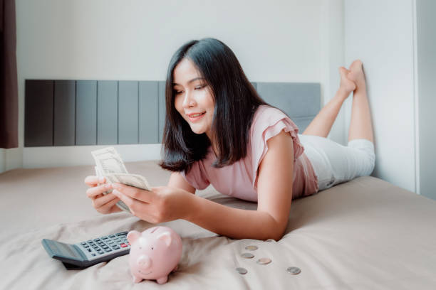 portrait de belle femme est calcul d'économie d'argent tout en se trouvant sur sa chambre à coucher. concept de gestion financière et d'investissement. - piggy bank currency savings finance photos et images de collection