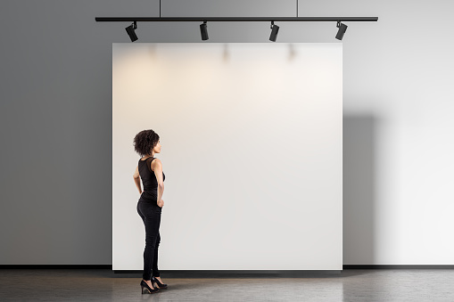 Beautiful African American woman wearing black clothes looking at blank gallery wall. Concept of advertising. Mock up