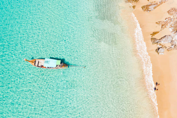 vista desde arriba, impresionante vista aérea de dos personas caminando en una hermosa playa tropical con arena blanca, agua cristalina turquesa y un barco de cola larga tradicional. banana beach, phuket, tailandia. - phi phi islands fotografías e imágenes de stock