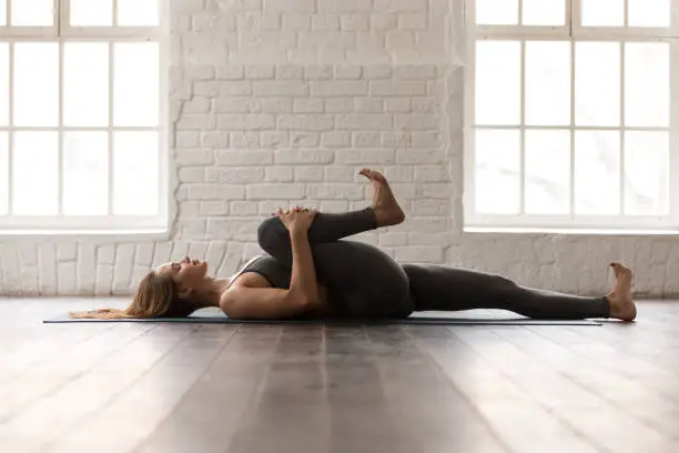 Calm woman with closed eyes practicing yoga, attractive girl in grey sportswear, pants and bra lying in Knees to Chest pose, doing Apanasana exercise, working out at home or in yoga studio