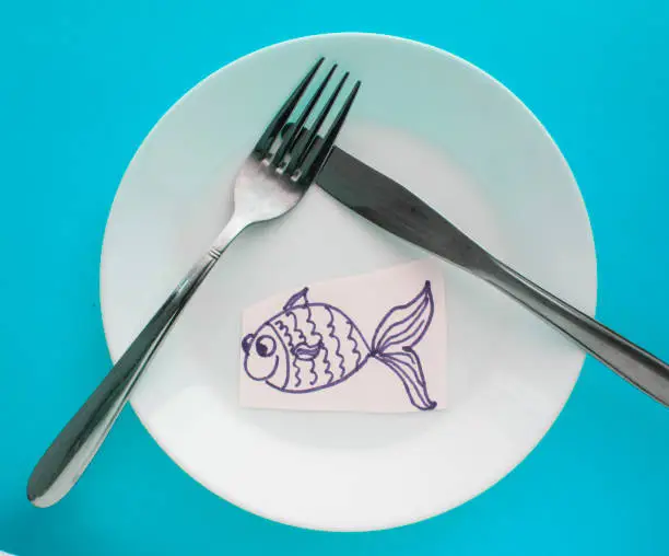 Photo of The celebration of April fool's Day, a Plate with a fork and knife and a paper fish on a blue background. Humor