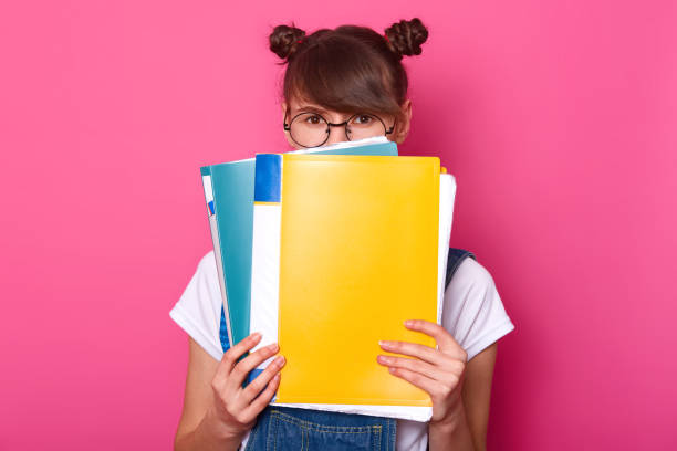 Image of surprised emotive european woman hides behind colourful paper folders, wears round black glasses, white t shirt, denim overalls, stands against pink background. Back to school concept. imagen de la mujer emotiva europea sorprendido se esconde detrás de las carpetas de papel colorido, lleva gafas redondas negro, camiseta blanca, overoles de mezclilla, se encuentra contra el fondo rosado. concepto de regreso a la escuela.