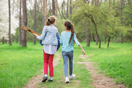 two happy young girls with guitar walking singing song along spring green park trail