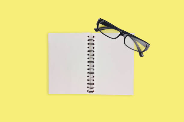 istock One transparent optical glasses in black plastic frame near paper notepad with blank sheets on table of yellow color. Top view. Business concept 1134881010