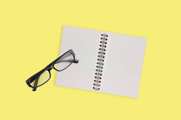 One transparent optical glasses in black plastic frame near paper notepad with blank sheets on table of yellow color. Top view. Business concept. Copy space for your text