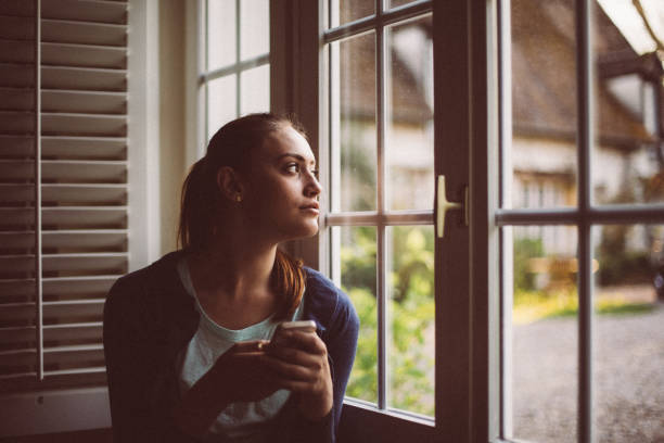 Cute girl looking through the window Woman at home texting on cell phone mesmerised stock pictures, royalty-free photos & images