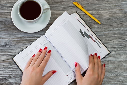 Woman's hands turning a page in a notebook. Cup of tea and a pen are on the background. Top view photo