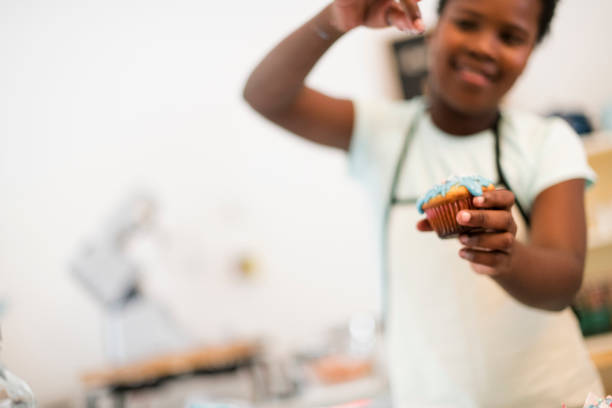 Young African girl decorating cupcakes with sprinkles, selective focus on cupcake.